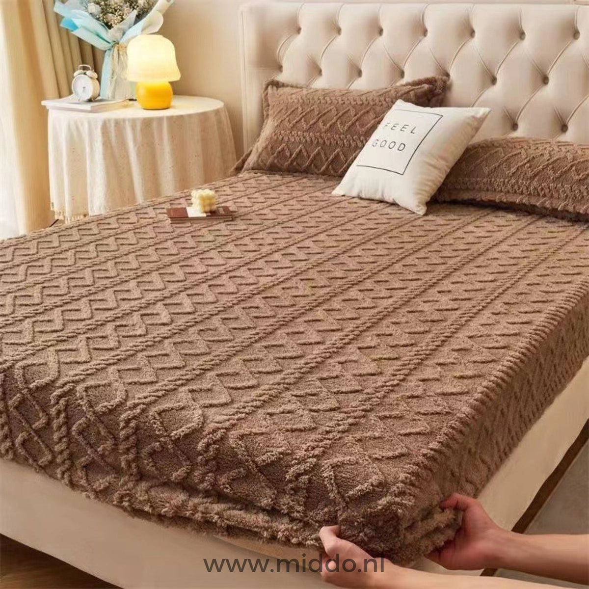 Deux mains tirant un drap-housse en polaire taupe sur l'angle du matelas dans une chambre avec tête de lit beige