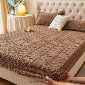 Deux mains tirant un drap-housse en polaire taupe sur l'angle du matelas dans une chambre avec tête de lit beige